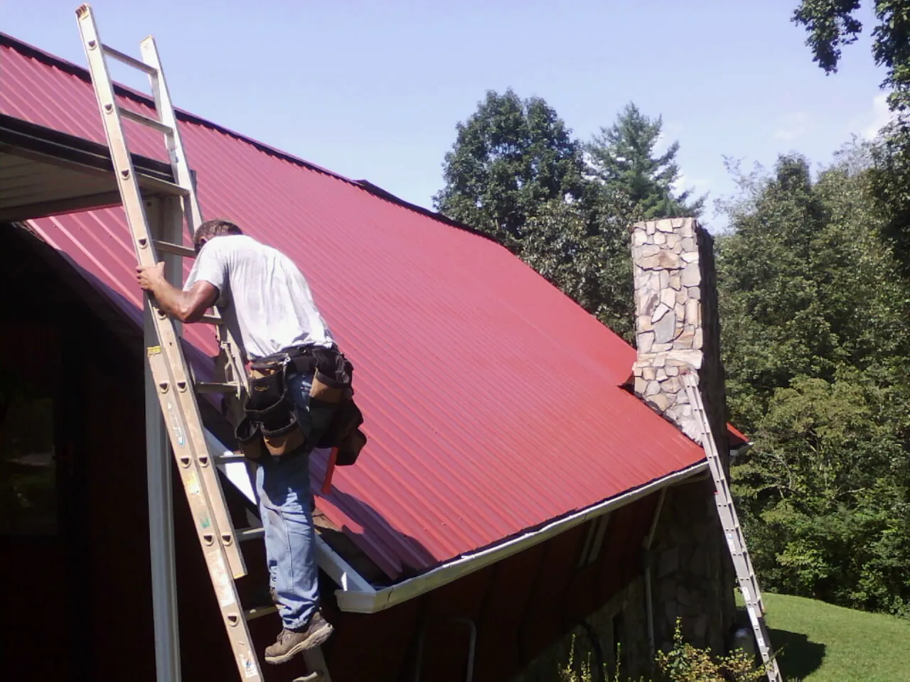 Roofer on a ladder beside a red metal roof and stone chimney — Soffit & Fascia Repair by Roofing Brothers in Lily